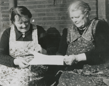 876863 Portret van twee onbekende vrouwen die een kinderhemdje bekijken, zittend aan een onbekende straat in Wijk C te ...